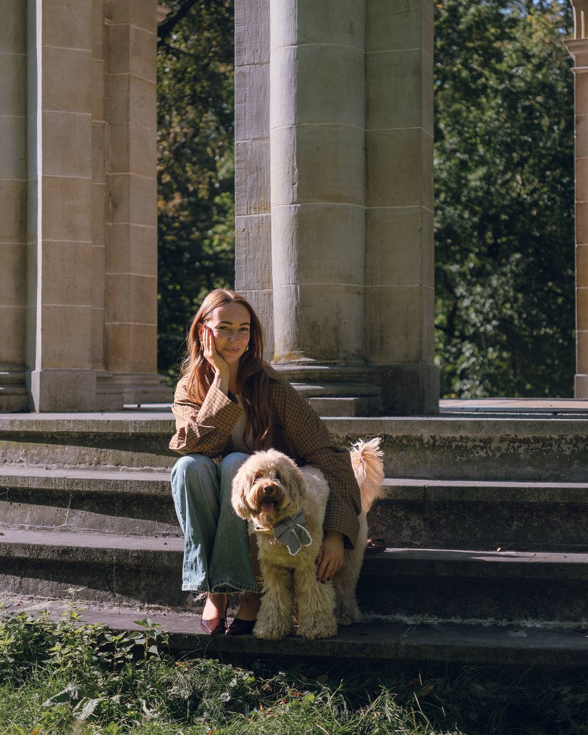 A woman sits on stone steps in front of large classical columns, holding a fluffy cream-colored dog. She is wearing a brown knit sweater and light blue jeans.
