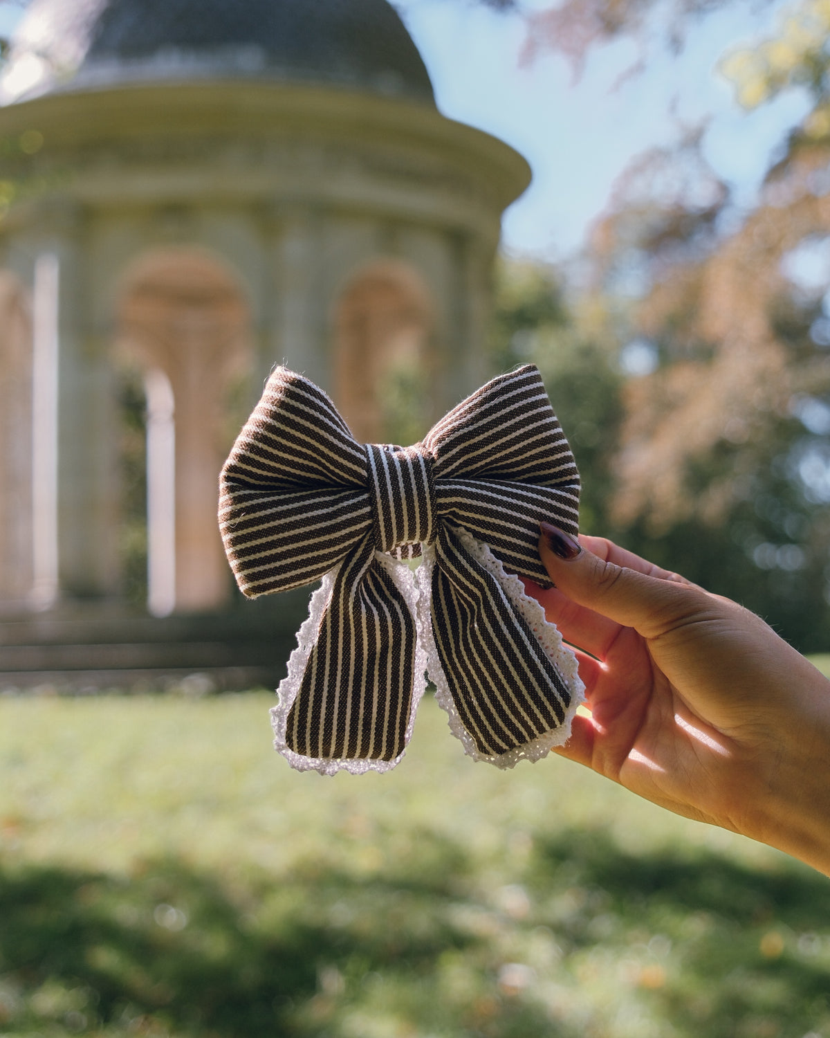 A hand holds a striped fabric bow with white lace trim in front of a blurred outdoor background featuring a classical stone gazebo and green grass.