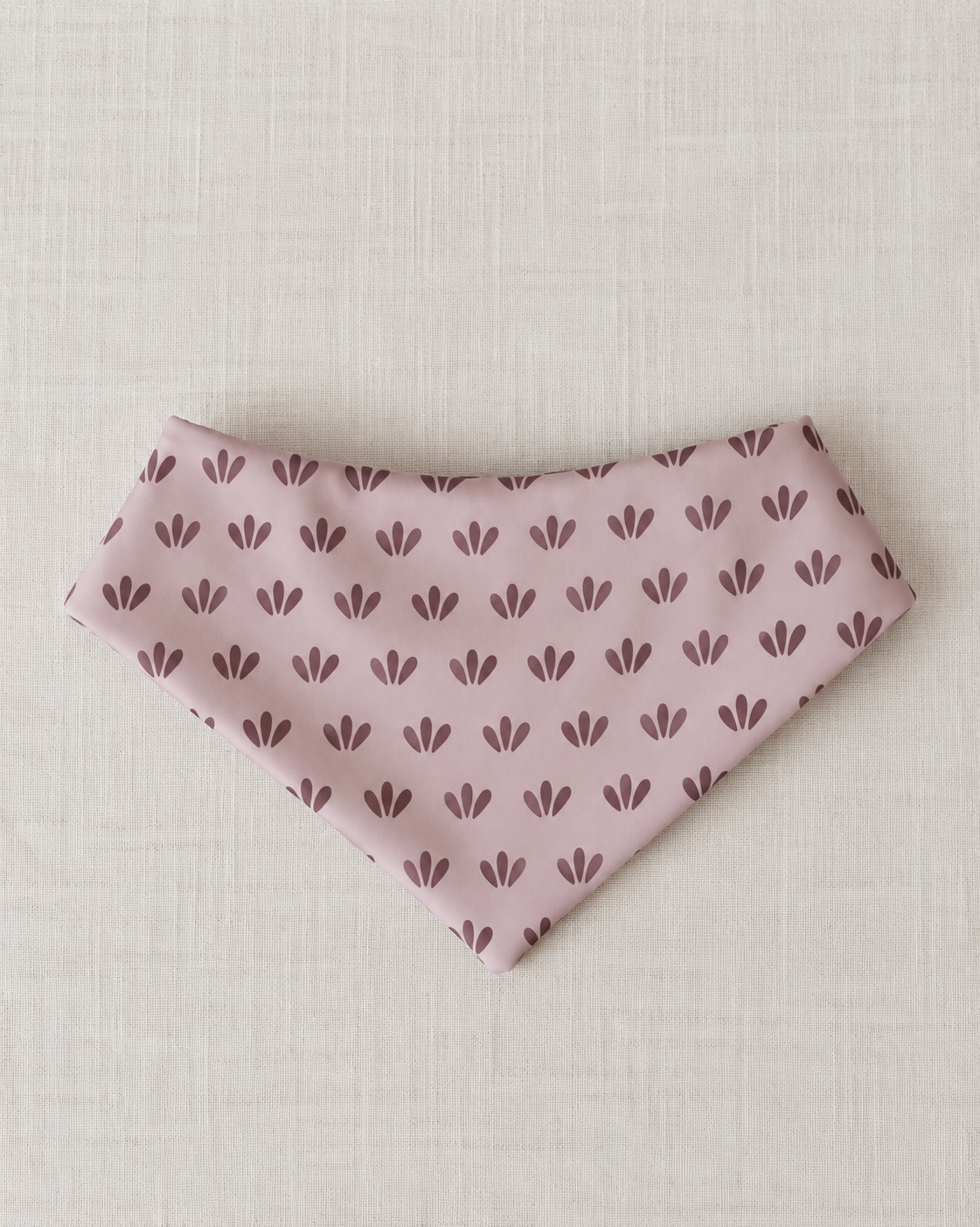 A light pink triangular bandana or pet bandana with a repeating dark plum leaf pattern, laid flat on a textured beige linen background.
