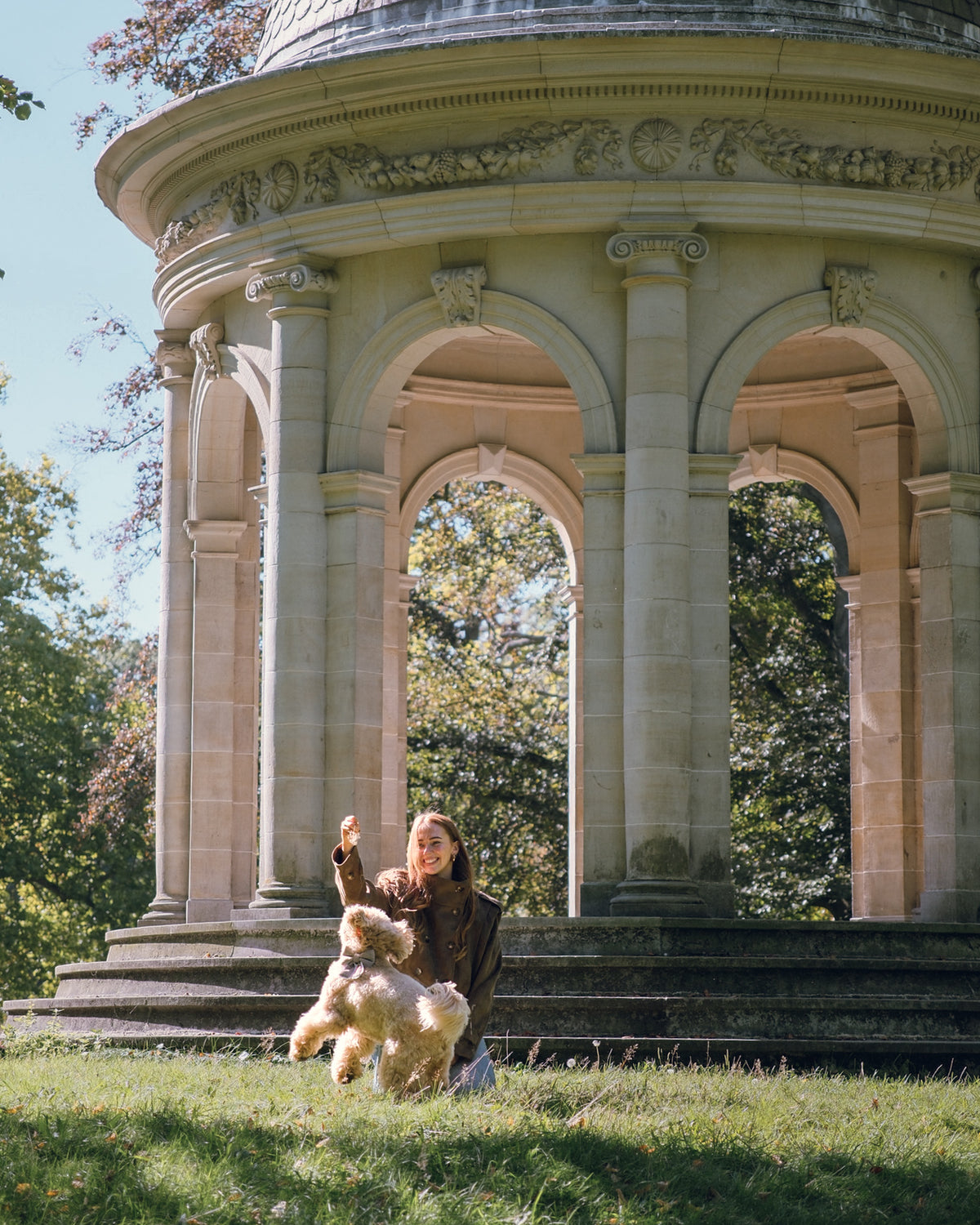 A woman kneels on a grassy lawn in front of a large stone rotunda, holding a fluffy cream-colored dog. The scene is outdoors with trees in the background and bright natural sunlight.