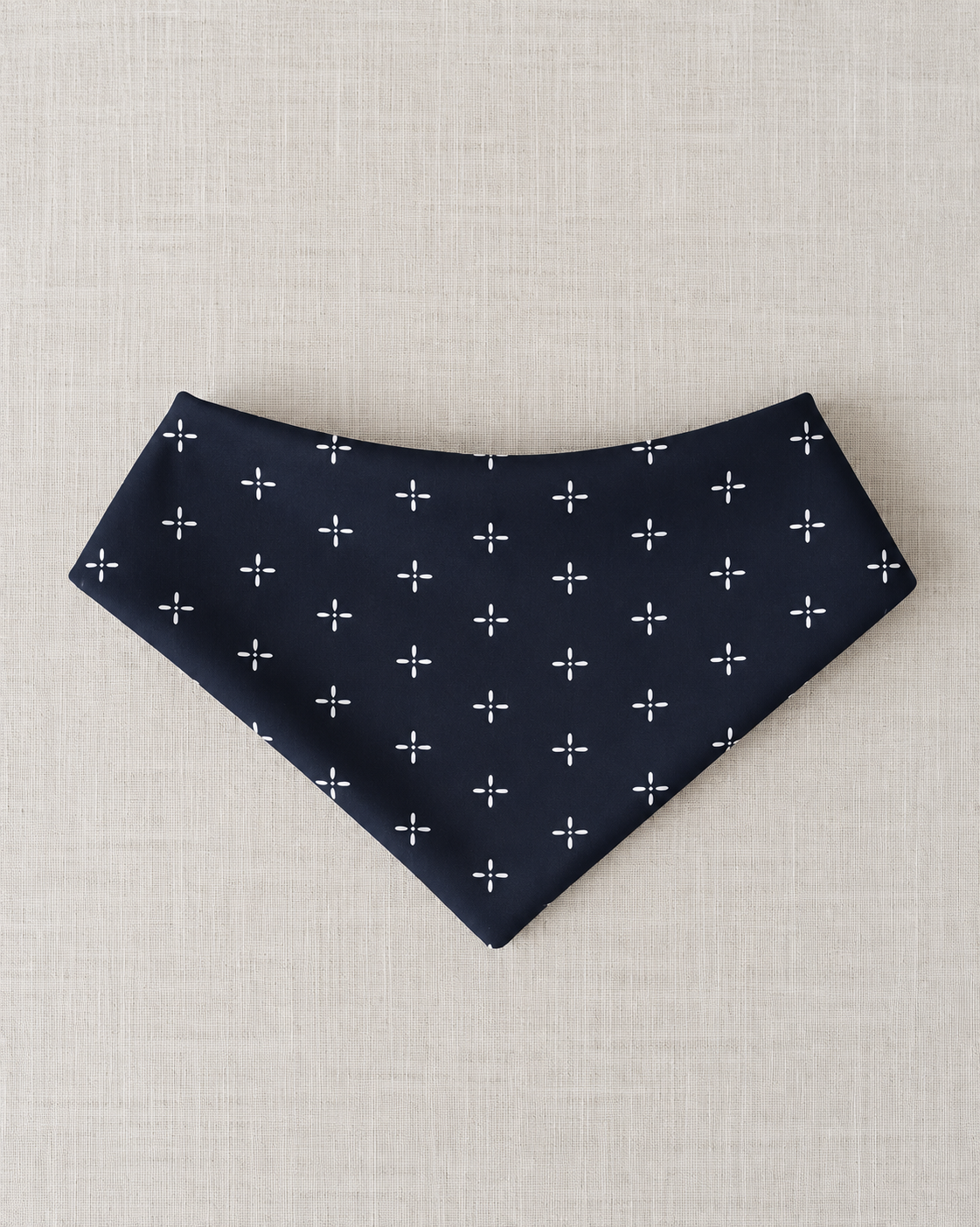 A navy blue triangular bandana or pet bandana with a white cross pattern, laid flat on a textured beige linen background.